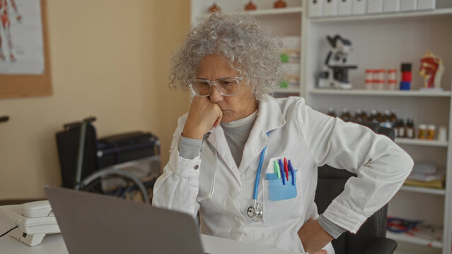 Woman doctor with grey hair thoughtfully looking at a laptop in a clinic office surrounded by medical equipment and bookshelves
