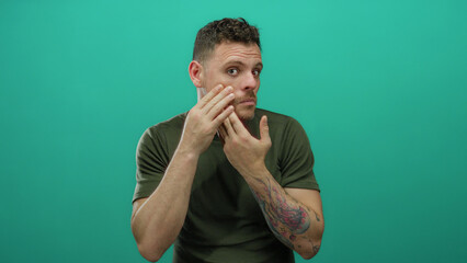 Young man with tattoos touching face against an isolated green background wearing casual clothing...