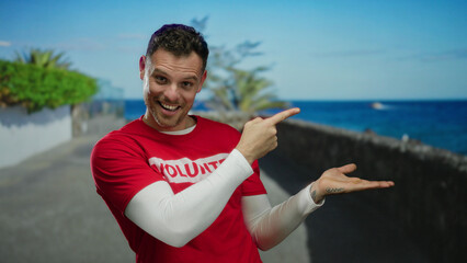 Young man in red shirt smiling and pointing with ocean view behind him along promenade with bright...