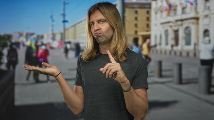 Man wearing a gray shirt presenting open palm while finger pointing upward on a sunny street; doubt.