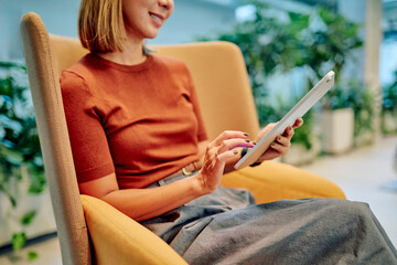 Smiling businesswoman using digital tablet while sitting in comfortable armchair in modern office lobby, working remotely