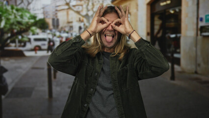 Fototapeta premium Blond man with long hair making hand binoculars gesture and smiling on busy street; curiosity discovery.