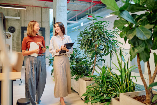 Two businesswomen walking through a vibrant green office, discussing work while holding tablets, exemplifying collaboration and teamwork