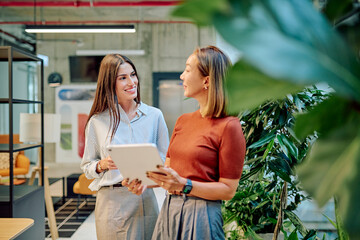 Two businesswomen are standing in a modern office, using a tablet and discussing, surrounded by plants