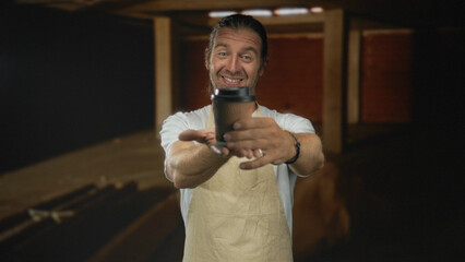 Blond man wearing apron extends coffee cup with both hands in building interior; friendly greeting...