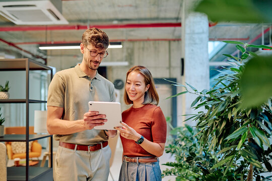 Two colleagues smiling and using digital tablet in modern office with plants, discussing project and sharing ideas