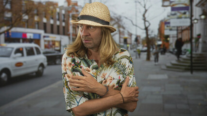 Blond tourist man hugs his shoulders wearing a straw hat and floral shirt on a street; restlessness...