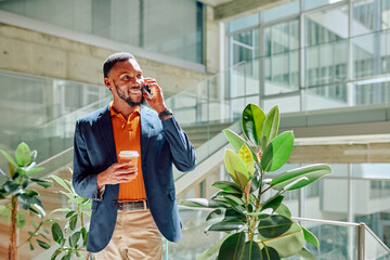 Young manager walking in office hallway, talking on smartphone and holding takeaway coffee cup,...