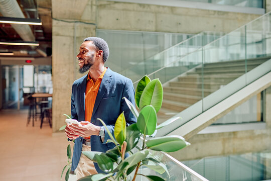 Young manager standing confidently in a corporate building hall, using a smartphone while surrounded by a vibrant green plant