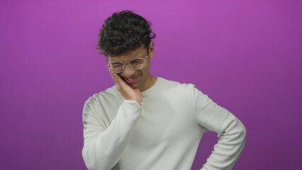 Man gently rubbing cheek in vivid purple studio set with glasses and knit sweater showing...