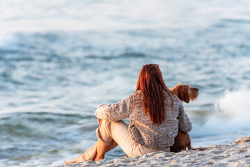 A red-haired woman sits on a sandy beach facing the sea, accompanied by a ginger dog. Baltic Sea, Poland.