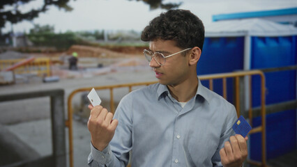 Young hispanic man frowning holds blue creditcard and white plastic card at outdoor construction...