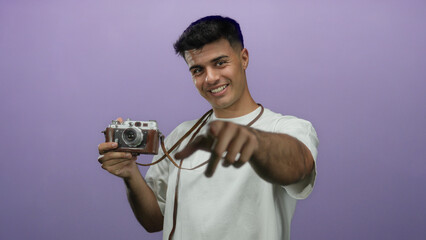 Smiling man holding vintage camera points towards viewer against purple background showcasing...