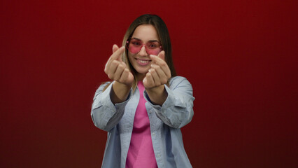 Woman making heart gesture wearing heart-shaped glasses with confident smile over red background isolated joyful female fashion pop art concept