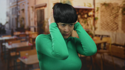 Woman in green shirt covering ears standing on restaurant terrace with outdoor seating in the...