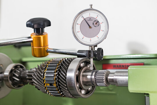 Gearbox shaft run-out measuring by dial indicator in holder on white background. Closeup a test probe and transmission gear mechanism with ball bearing and synchronizer rings clamped in lathe machine.