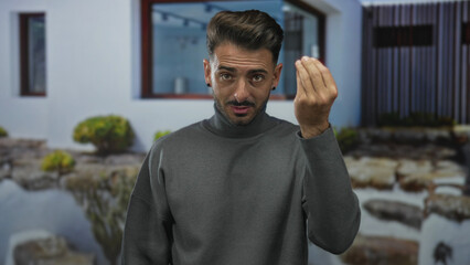 Young hispanic man gestures with pinched hand motion at modern house entrance on quiet city street;...