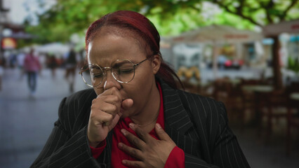 Woman coughing in busy city street among blurred pedestrians, hand covering mouth and chest;...