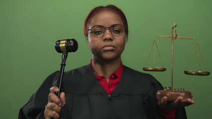 Serious african american judge holds gavel and scales in green studio setting with eyes closed;...