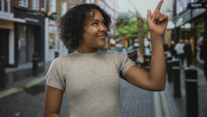 Young smiling woman standing pointing finger upward on busy wet city street lined with shops;...
