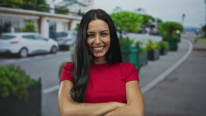 Fototapeta premium Woman in red shirt smiling on city street with cars in background and urban scenery, outdoor environment highlighting youthful energy and hispanic style.