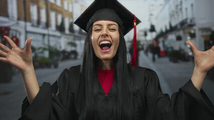 Young hispanic woman in graduation cap and gown smiling joyfully outdoors in a city street,...