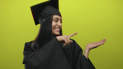 Woman in graduation gown pointing to her palm over an isolated yellow background, embodying joy and...