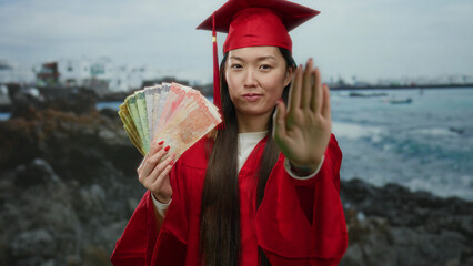Woman in red graduation attire holding philippine peso banknotes near seaside with a serious...