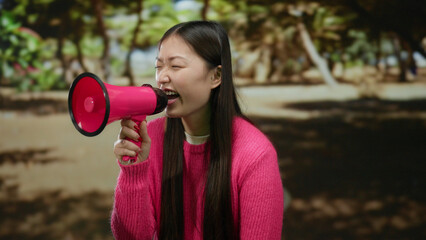 Woman holding megaphone outdoors in park wearing pink sweater with trees and blurred background...
