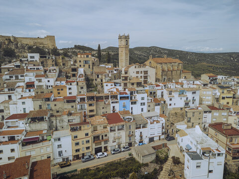 Aerial view of terracotta rooftops cascading down the hillside towards the towering church spire and ancient castle ruins, Cervera del Maestre, Valencian Community, Spain.