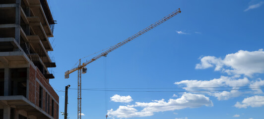 Wide view of a yellow tower crane next to a concrete building under construction, set against a blue sky with scattered clouds.