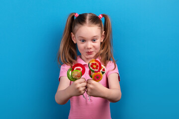 Surprised and excited young girl in a pink shirt holding colorful fruit-shaped lollipops against a vibrant blue background.