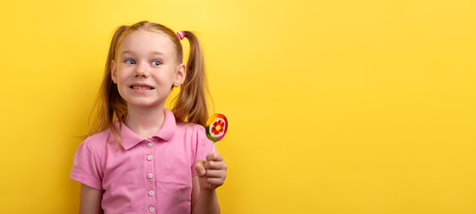 Amused young girl in a pink shirt holding a colorful lollipop and making a wide smile against a yellow background.