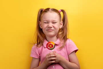 Young girl in pink shirt making a funny grimace while holding a colorful lollipop against a vibrant yellow background.