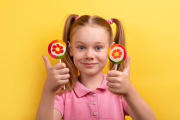 Happy young girl with pigtails holding two colorful lollipops and giving a thumbs up in front of a bright yellow background.