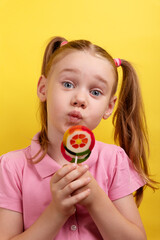  Cute young girl with pigtails playfully pouting while holding a colorful lollipop in front of a bright yellow background.