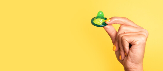 Hand with yellow-painted nails holding a green lubricated condom by the rim against a solid yellow background.