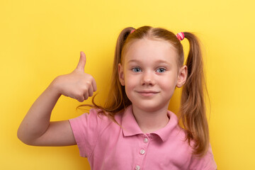 Cheerful young girl in pink shirt giving thumbs up gesture against vibrant yellow background, symbolizing approval and positivity.