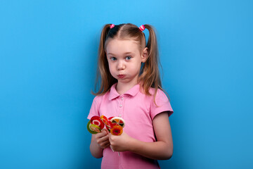  Cute girl in a pink shirt puffing her cheeks while holding colorful lollipops against a bright blue background.