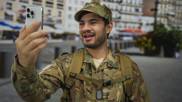 Man in military uniform holds smartphone for a video call on a busy urban street lined with apartment buildings; connection communication camaraderie service. - Powered by Adobe