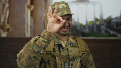 Young hispanic man in camouflage military uniform shows ok sign with hand in ancient building...