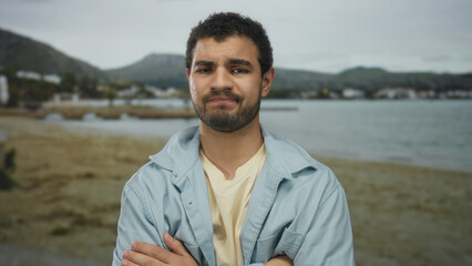Young hispanic man standing with crossed arms and furrowed brow wearing light blue shirt at beach; uncertainty.