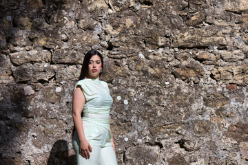 A beautiful Spanish teenager with long dark hair poses like a model on the green grass next to a stone wall of a ruined castle on the outskirts of the city. Concept beauty and fashion.