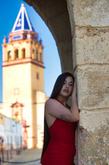 A beautiful teenage girl with long dark hair dressed in red leaning under a Roman archway on a street in a typical Andalusia town. In the background is a church tower.