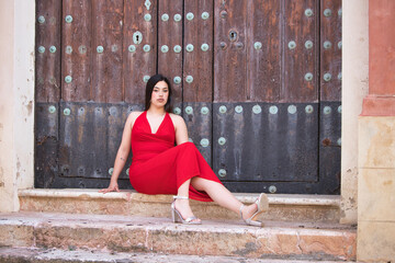 A pretty teenage girl with long dark hair, dressed in red and wearing high heels, sitting on the steps of a church and leaning against the wooden door.