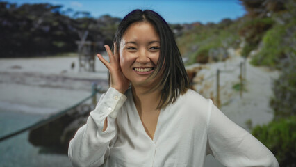 Woman in park gestures happily with thumb up and hand to ear, dressed in white shirt, enjoys sunny...