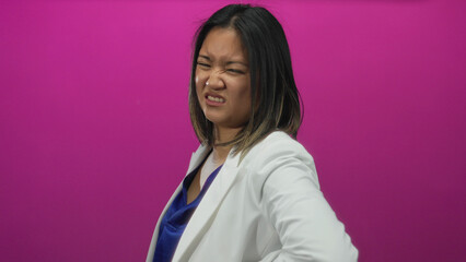 Chinese woman in white blazer looks pained, holding her lower back against a vibrant pink...