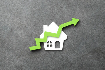 A white house model with a green upward arrow, symbolizing rising property value on a stone...