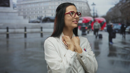 Fototapeta premium Woman in a lab coat stands pensively on a london street, surrounded by red buses, showcasing her hispanic heritage amid an urban backdrop.