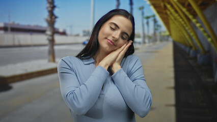 Young hispanic woman stretches and mimics sleep outdoors on a sunny street, wearing a blue shirt, enjoying her day underneath palm trees and clear blue skies.